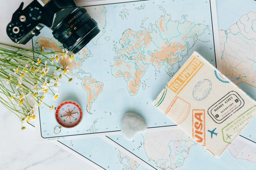 Vue du dessus d'une carte du monde étalée sur une table, entourée d'un appareil photo vintage noir, d'un carnet de voyage en tissu imprimé de tampons de visas, d'une boussole rouge et d'un petit bouquet de petites fleurs de camomille.